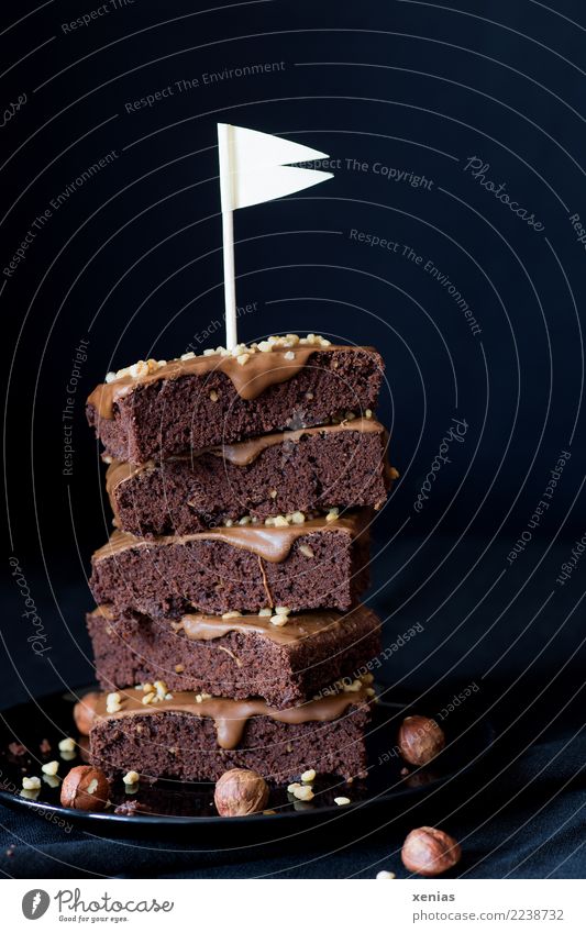 aufgetürmte Stücke Schokoladenkuchen mit Haselnusscreme und weißer Fahne vor dunklem Hintergrund Lebensmittel Kuchen Brownie Creme Nusscreme Krokant Bioprodukte