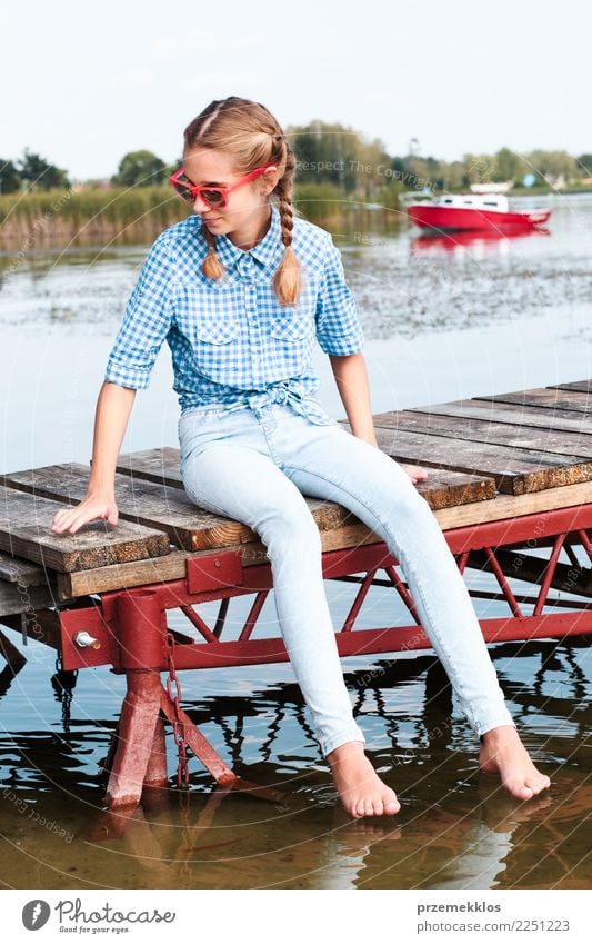 Junges Mädchen sitzt auf dem Steg über dem See und taucht die Füße ins Wasser an einem sonnigen Sommertag Lifestyle Freude Glück Erholung Freizeit & Hobby