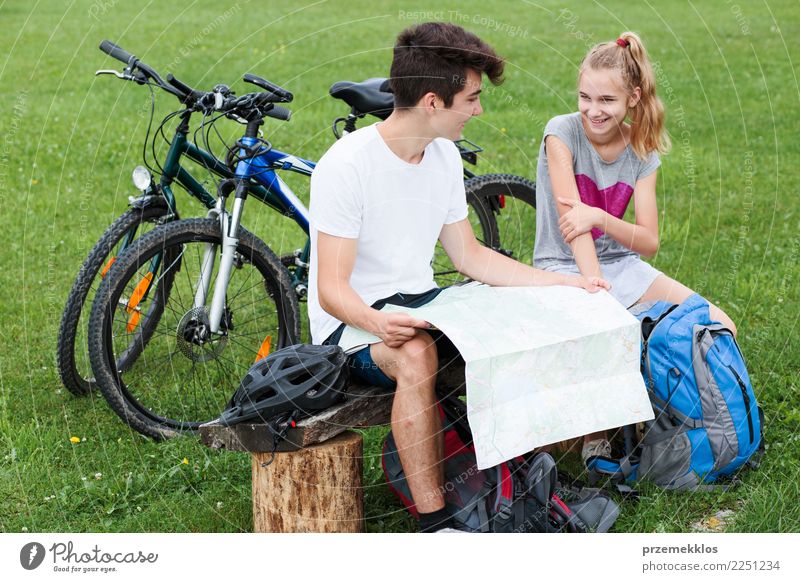 Junger lächelnder Junge und Mädchen machen eine Pause auf einer Bank sitzend während eines Fahrradausflugs und planen einen weiteren Teil der Reise mit einer Karte, ausgestattet mit Helmen, Rucksäcken und Fahrrädern