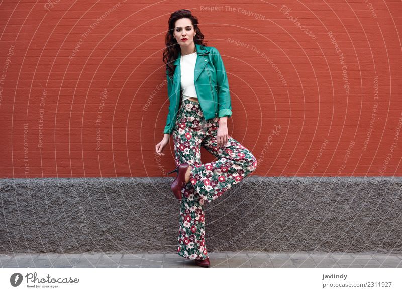 Junge brünette Frau, Modell der Mode, trägt grüne moderne Jacke und Blume Hose auf rote Wand Lifestyle Stil schön Haare & Frisuren Gesicht Mensch feminin