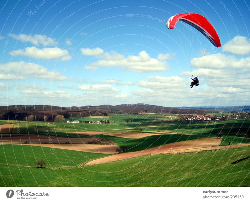 Paraglider mit rotem Gleitschirm fliegt über grüne Wiesen und Felder vor Himmel mit Wolken paraglider Gleitschirmfliegen Paragliding Abenteuer Freiheit Sportler