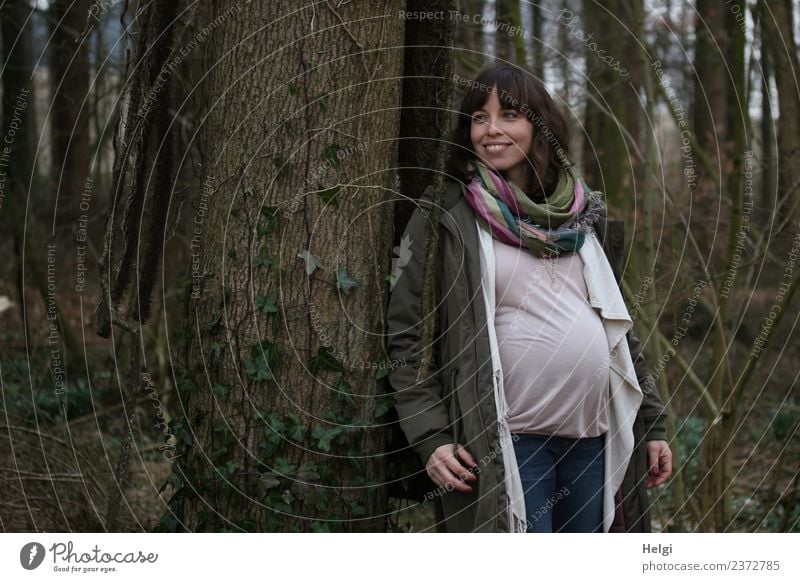 junge schwangere Frau steht lächelnd an einem Baum Mensch feminin Junge Frau Jugendliche Erwachsene 1 30-45 Jahre Umwelt Natur Wald Bekleidung T-Shirt Mantel