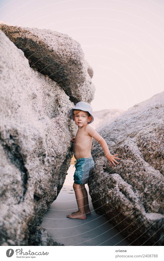 kleiner kaukasischer Junge klettert zwischen großen Felsen am Strand. Sommer Klettern Bergsteigen Kind Kleinkind Kindheit Wärme Badehose Hut blond Streifen