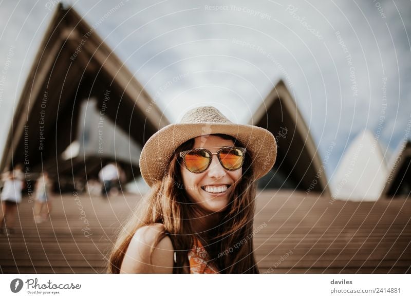 Reizende Frau mit Hut und Sonnenbrille, die in die Kamera schaut, während sie das Opernhaus in Sydney, Australien, besucht. Lifestyle Freude