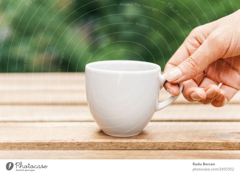 Frau Hand, die eine Tasse Kaffee von einem Holztisch im Garten aufzieht. Tisch Hintergrundbild grün Frühstück Morgen Café Licht weiß trinken Espresso