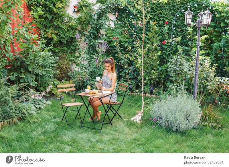Junge und attraktive Frau beim Frühstück am Morgen im grünen Garten mit französischem Croissant, Donuts, Kaffeetasse, Orangensaft, Tablette und Notizbuch auf Holztisch.