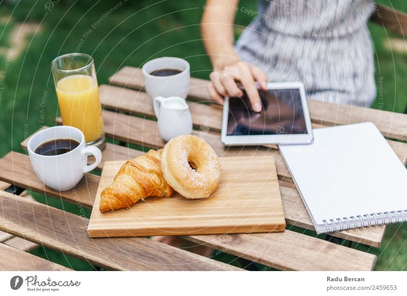 Junge und attraktive Frau beim Frühstück am Morgen im grünen Garten mit französischem Croissant, Donuts, Kaffeetasse, Orangensaft, Tablette und Notizbuch auf Holztisch.