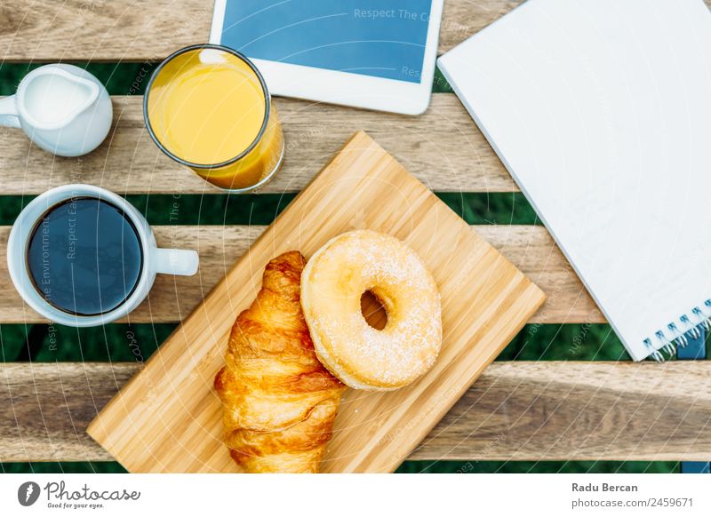 Frühstück im Grünen Garten mit französischem Croissant, Donuts, Kaffeetasse, Orangensaft, Tablette und Notizen Buch auf Holztisch Tisch Hintergrundbild Morgen