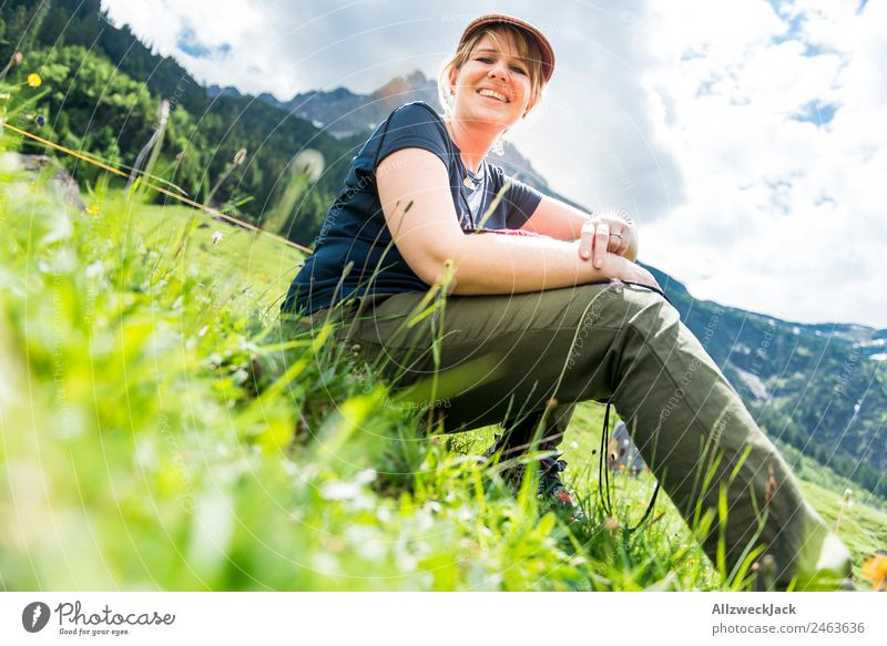 junge Frau sitzt im Gras mit Bergen im Hintergrund Tag Schönes Wetter Wolken Natur grün Baum Wald Berge u. Gebirge Idylle Ferien & Urlaub & Reisen