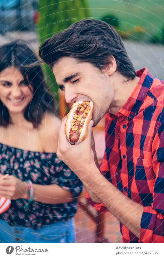 Junger Mann isst Hot Dog und Frau lacht im Hintergrund. Wurstwaren Brot Brötchen Mittagessen Fastfood Lifestyle Freude Glück Sommer Erwachsene Freundschaft Hand