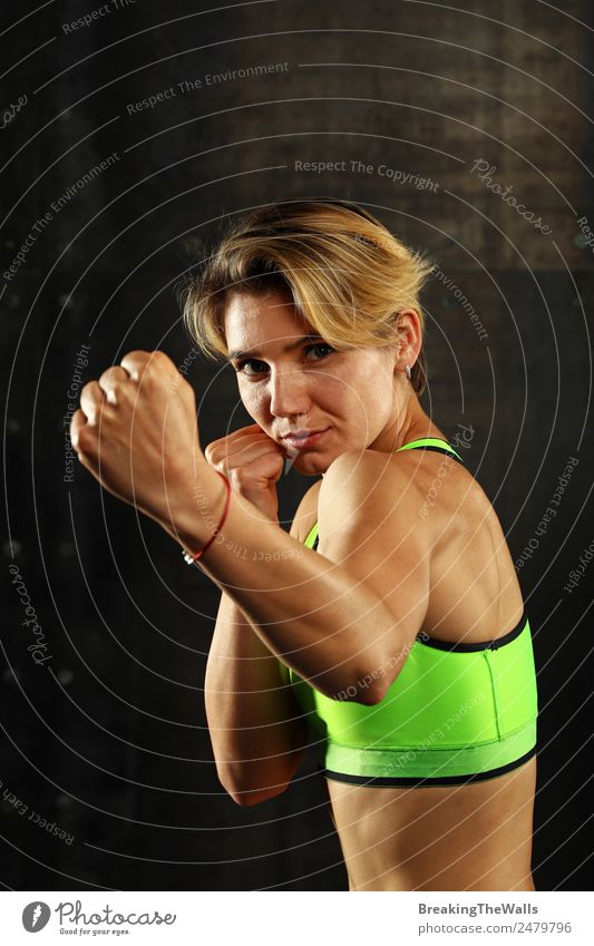 Nahaufnahme Frontalporträt einer jungen sportlichen Frau in Sportkleidung in einer Turnhalle vor dunklem Hintergrund, stehend in Boxhaltung mit Händen und Fäusten, Blick in die Kamera