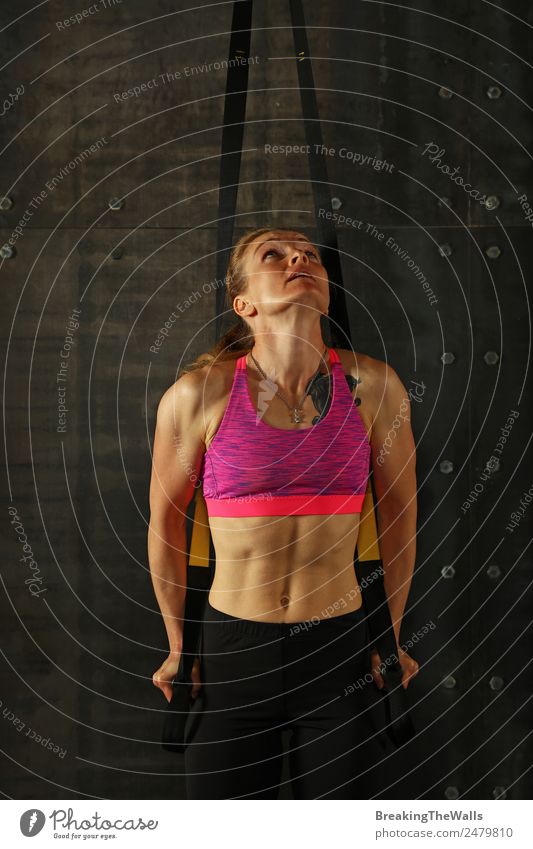 Eine junge, sportliche Frau mittleren Alters beim Crossfit-Training, die mit Trx-Suspension-Fitnessbändern vor dunklem Hintergrund trainiert, Vorderansicht, Blick nach oben