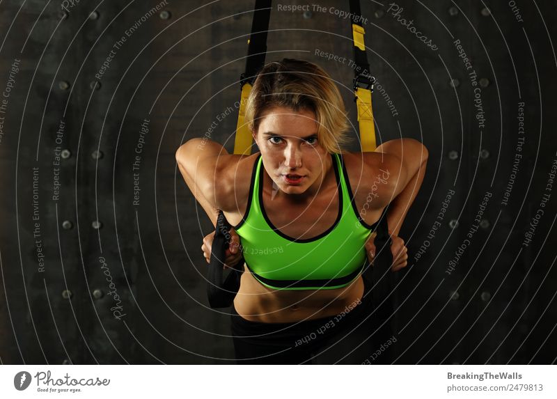 Eine junge sportliche Frau beim Crossfit-Training, die mit Trx-Suspension-Fitnessbändern vor dunklem Hintergrund trainiert, Vorderansicht, Blick in die Kamera