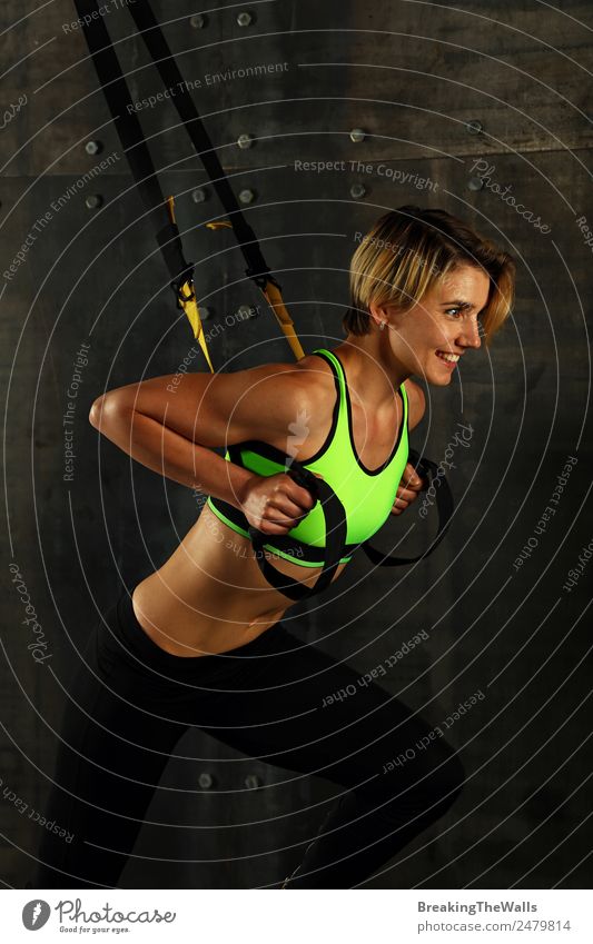 Seitenprofilansicht Porträt einer jungen sportlichen Frau beim Crossfit-Training, die mit Trx-Suspension-Fitnessbändern vor dunklem Hintergrund trainiert und wegschaut