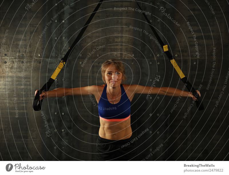 Eine junge, sportliche Frau mittleren Alters beim Crossfit-Training, die mit Trx-Suspension-Fitnessbändern vor dunklem Hintergrund trainiert, Vorderansicht, Blick in die Kamera