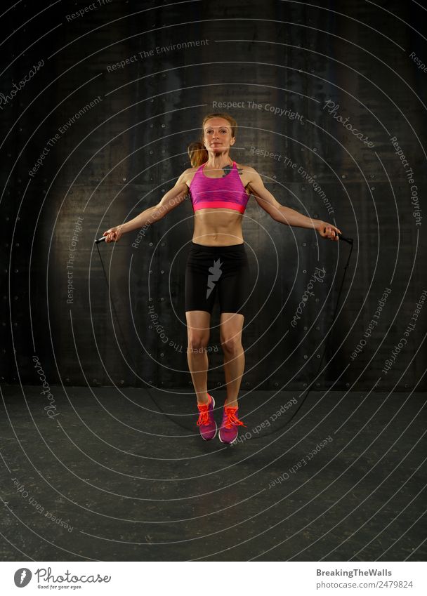 Eine junge, sportliche Frau mittleren Alters, die trainiert, mit Fitness-Springseil über dunklem Hintergrund, in voller Länge Vorderansicht, Blick in die Kamera