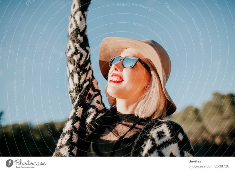 Nahaufnahme eines Porträts einer glücklichen Frau mit Sonnenbrille und Hut, die den Sonnenuntergang in der Natur genießt und den Arm hebt. Lifestyle Stil