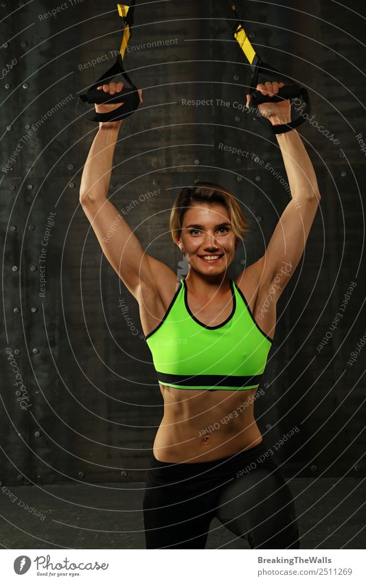 Eine junge sportliche Frau beim Crossfit-Training, die mit Trx-Suspension-Fitnessbändern vor dunklem Hintergrund trainiert, Vorderansicht, Blick in die Kamera