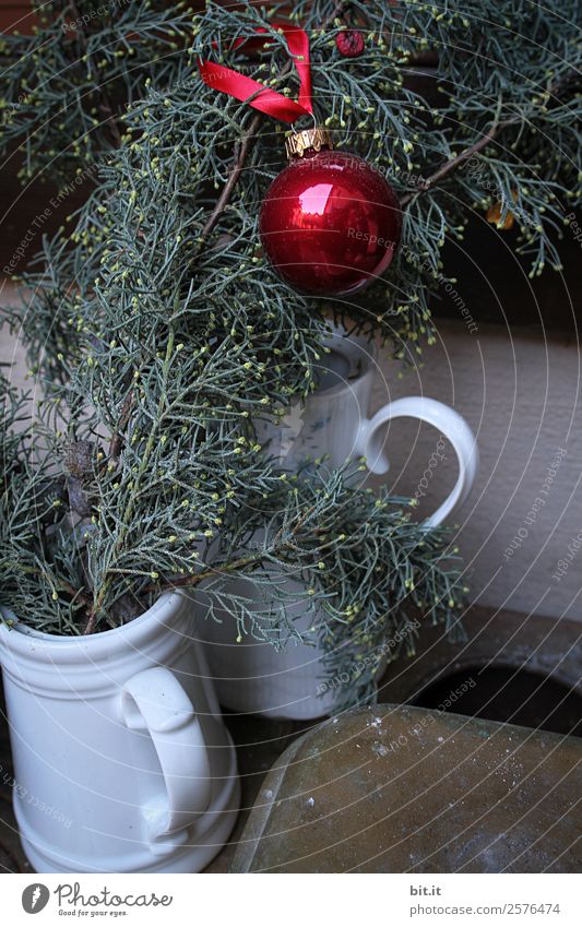 Nostalgische Weihnachtsdekoration mit zarten Tannenzweigen, roter Kugel und Vasen auf altem Tisch. Rote Weihnachtskugel hängt glänzend an Zweigen. Leuchtende Christbaumkugel aufgehängt an Ästen in alten weissen, nostalgischen Kannen aus Porzellan zuhause.