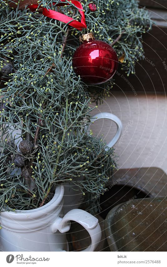 Nostalgische Weihnachtsdekoration mit zarten Tannenzweigen, roter Kugel und Vasen auf altem Tisch. Rote Weihnachtskugel hängt glänzend an Zweigen. Leuchtende Christbaumkugel aufgehängt an Ästen in alten weissen, nostalgischen Kannen aus Porzellan zuhause.