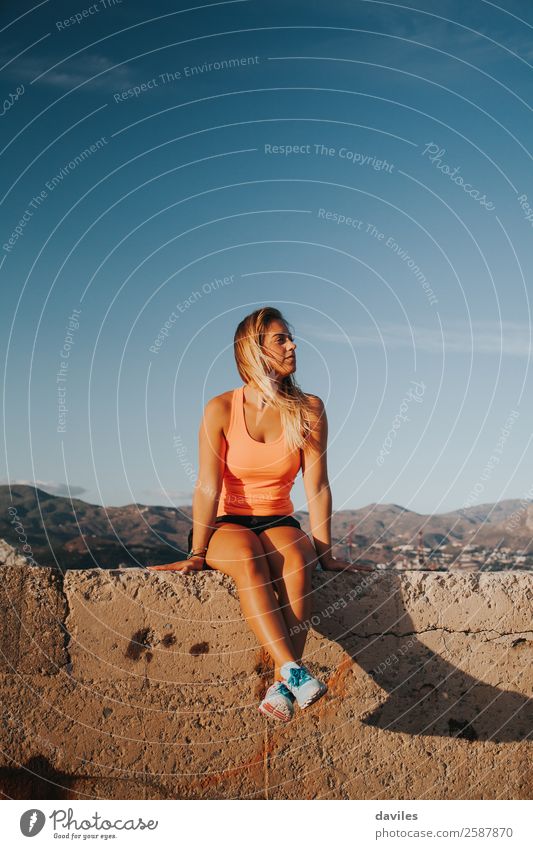 Schöne Frau in Sportkleidung, die bei Sonnenuntergang auf einer Betonmauer im Freien sitzt. Lifestyle Berge u. Gebirge Fitness Sport-Training Mensch feminin