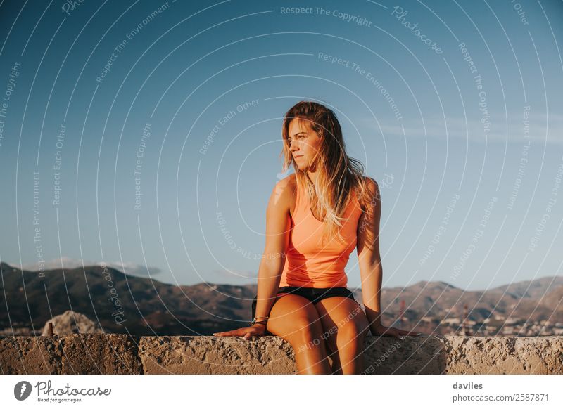 Schöne Frau in Sportkleidung, die bei Sonnenuntergang auf einer Betonmauer im Freien sitzt. Lifestyle Berge u. Gebirge Fitness Sport-Training Mensch feminin