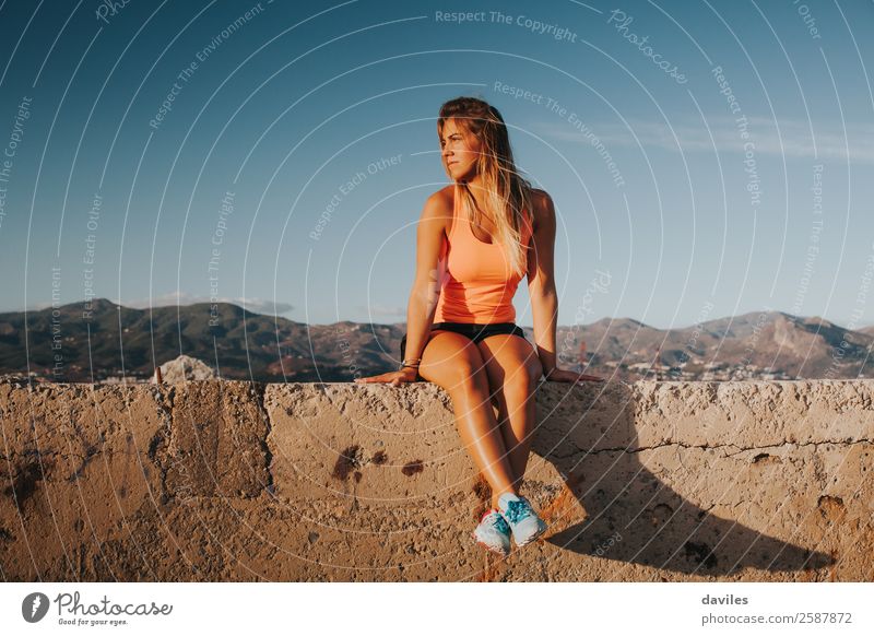 Schöne Frau in Sportkleidung, die bei Sonnenuntergang auf einer Betonmauer im Freien sitzt. Lifestyle Körper sportlich Wellness Wohlgefühl Berge u. Gebirge