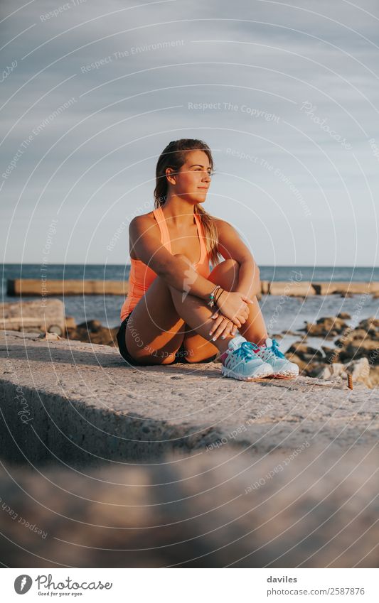 Schöne Frau in Sportkleidung, die bei Sonnenuntergang auf einer Betonmauer im Freien sitzt. Lifestyle Freude Wellness Erholung Meer Fitness Sport-Training