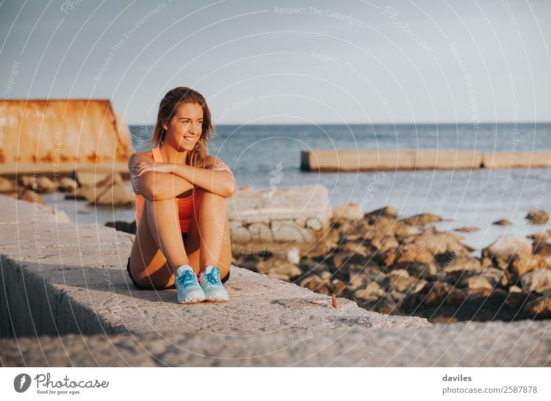 Lächelnde Frau in Sportkleidung, die bei Sonnenuntergang auf einer Betonmauer im Freien sitzt. Lifestyle Freude Glück schön Körper Erholung Freizeit & Hobby