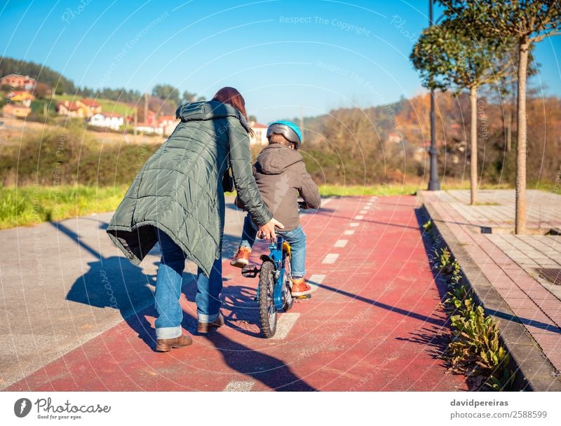 Rückansicht der Mutter, die ihrem Sohn das Fahrradfahren beibringt. Lifestyle Freude Glück Freizeit & Hobby Spielen Sonne Winter Sport Kind Schule Junge Frau