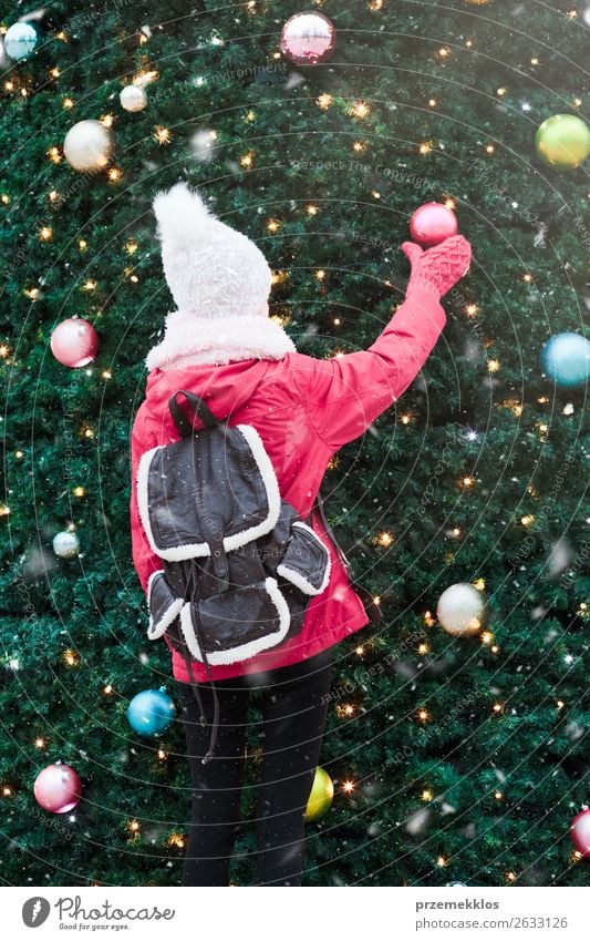 Junges Mädchen steht vor einem großen, mit bunten Kugeln geschmückten Weihnachtsbaum. Teenager Mädchen trägt warme Kleidung, Schal und Wollmütze an einem kalten Wintertag