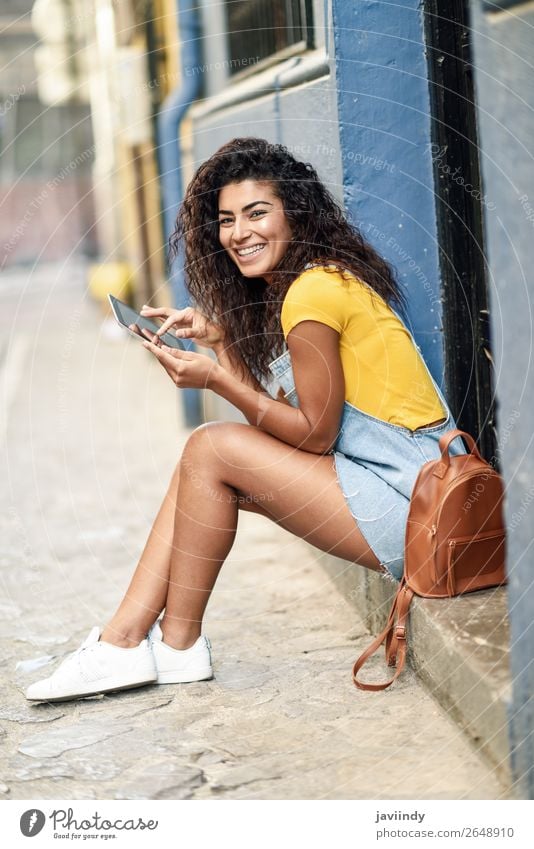 Fröhliche arabische Frau, die auf einer städtischen Stufe mit einem digitalen Tablett sitzt. Lifestyle Stil Glück schön Haare & Frisuren Tourismus