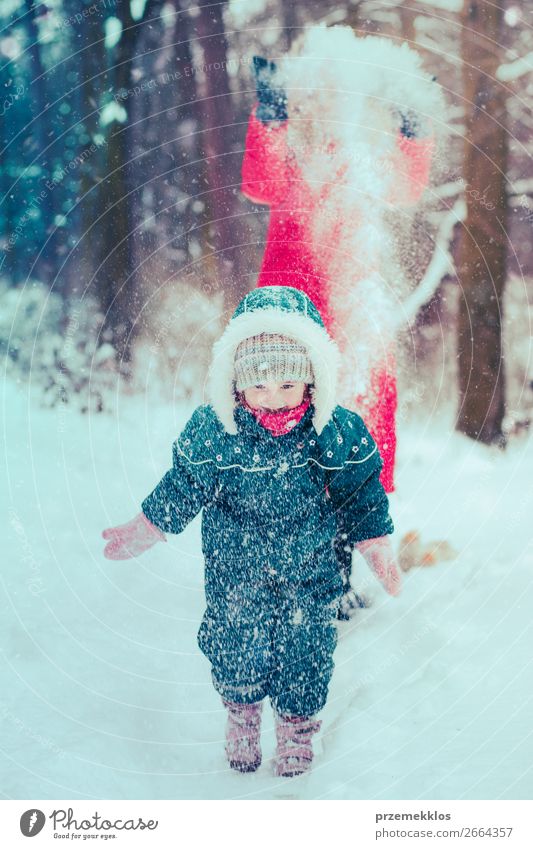 Mutter spielt mit ihrer kleinen Tochter im Freien an einem winterlichen Tag. Frau wirft Schnee auf ihr Kind. Familie verbringt Zeit zusammen und genießt die Winterzeit. Frau trägt roten Mantel und Wollmütze, Kleinkind trägt dunkelblauen Schneeanzug
