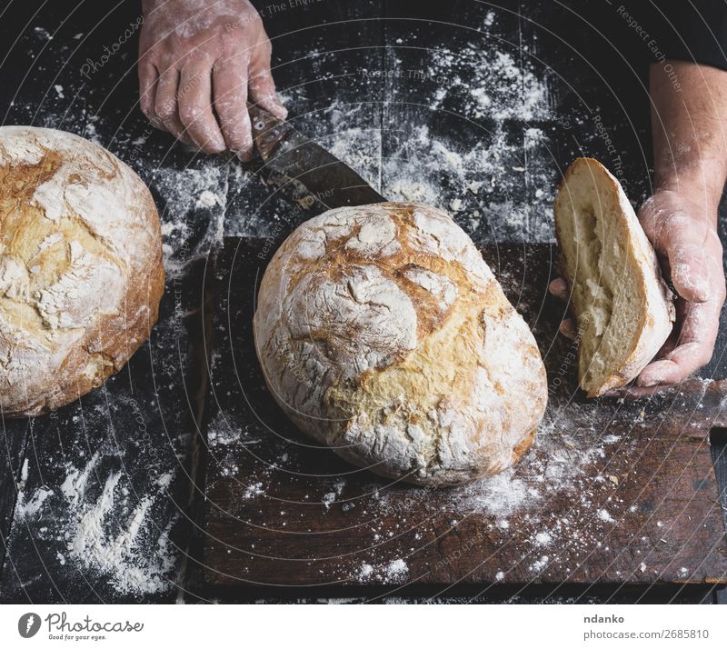 Mann schneidet gebackenes Rundbrot auf einem braunen Holzbrett. Brot Ernährung Tisch Küche Mensch Hand Finger machen dunkel frisch schwarz weiß Tradition ganz