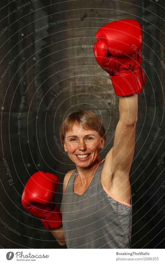 Frontalporträt einer jungen erwachsenen Frau in roten Boxhandschuhen mit der Geste des Siegers oder Champions in der Hand, lächelnd und in die Kamera blickend