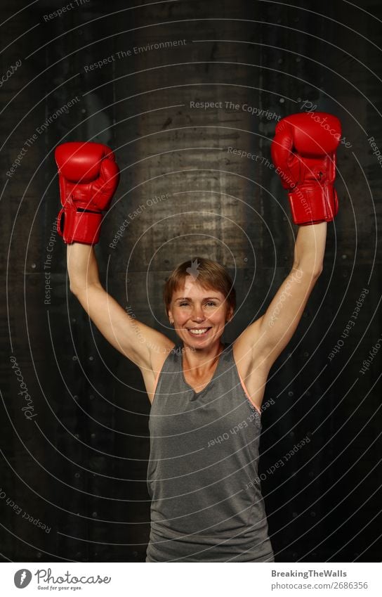 Frontalporträt einer jungen erwachsenen Frau in roten Boxhandschuhen mit zwei erhobenen Händen als Geste des Siegers oder Champions, lächelnd und in die Kamera blickend
