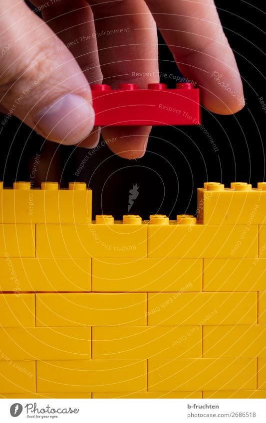 Spiel-Bausteine, gelbe Mauer, roter Baustein in der Hand Spielen Modellbau Hausbau Renovieren Handwerker Baustelle Finger Spielzeug Kunststoff
