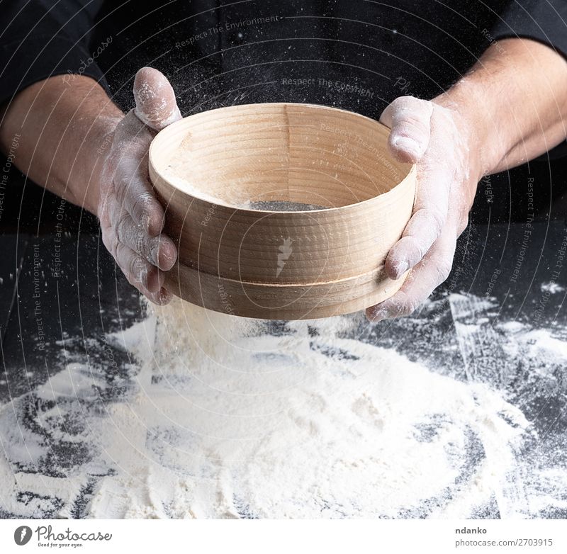 Koch in schwarzer Uniform hält in der Hand ein Sieb. Teigwaren Backwaren Brot Ernährung Tisch Küche Mensch Mann Erwachsene Holz Bewegung machen frisch weiß