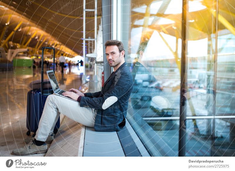 Mann, der am Flughafen sitzt und Laptop und Handy neben dem Fenster benutzt. Jugendliche Lächeln warten Etage Sonne Sonnenaufgang Telefon Kaukasier Sonnenlicht