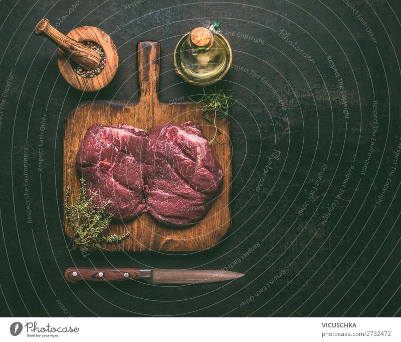 Rohes Marmor-Rindfleisch auf Holzschneidebrett mit Kräutern, Gewürzen, Öl und Messer auf dunklem, rustikalem Hintergrund, Draufsicht. Kopierfeld für Ihr Design oder Kochrezepte