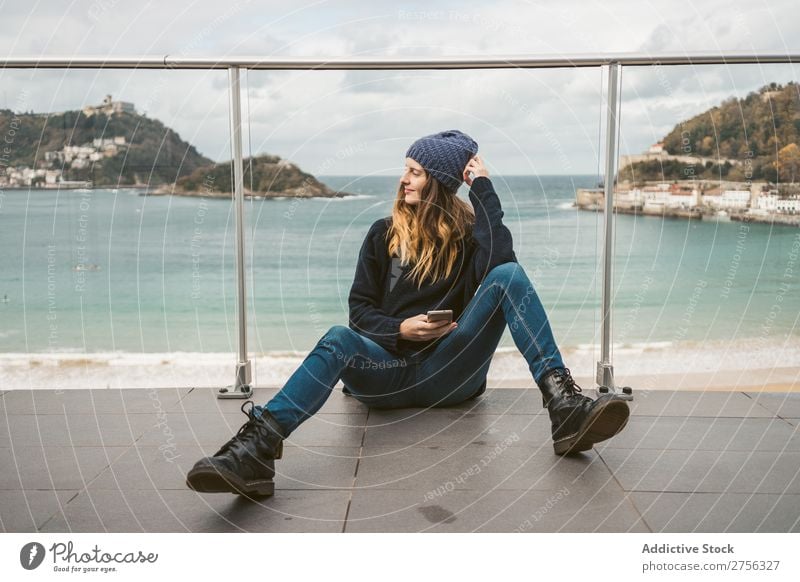 Frau sitzt mit Smartphone am Meer. Jugendliche Küste aufreizend Lächeln PDA sitzen Geländer benutzend hübsch attraktiv Hut Natur Wasser Ferien & Urlaub & Reisen