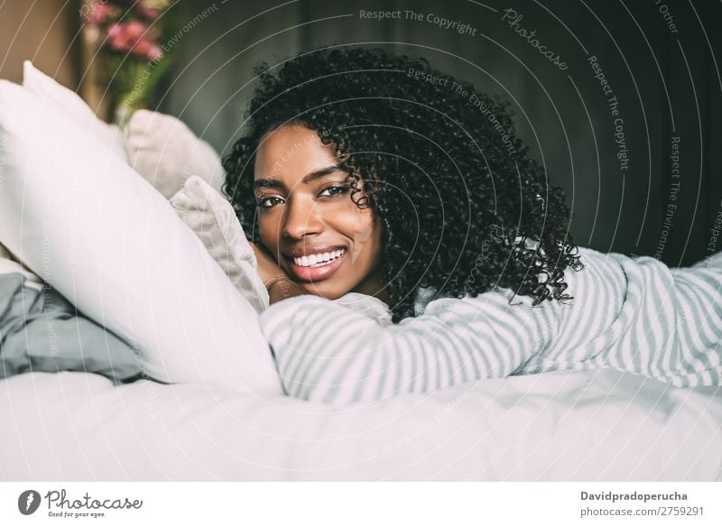 Nahaufnahme einer hübschen schwarzen Frau mit lockigem Haar, die lächelt und auf dem Bett liegt und auf die Kamera schaut. schwarze Frau Porträt lügen Lächeln