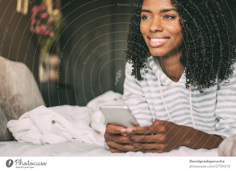 Nahaufnahme einer hübschen schwarzen Frau mit lockigem Haar, die lächelt und Telefon auf dem Bett benutzt, um wegzuschauen. schwarze Frau Porträt PDA Mobile