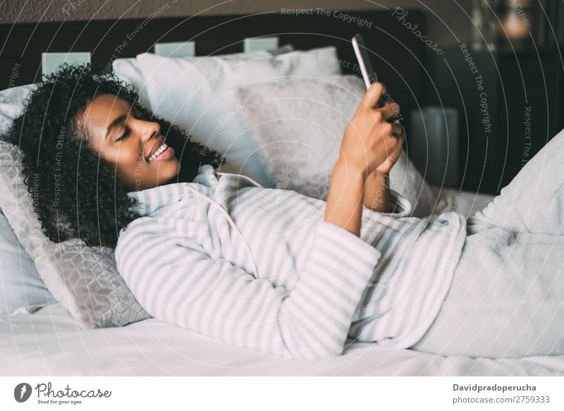 Nahaufnahme einer hübschen schwarzen Frau mit lockigem Haar, die lächelt und Telefon auf dem Bett benutzt, um wegzuschauen. schwarze Frau Porträt PDA Mobile