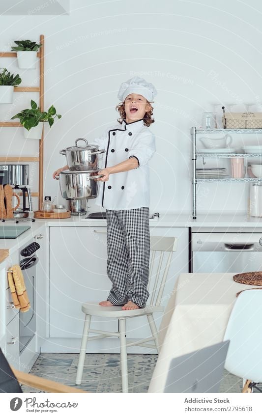 Lächelnder Junge mit Kochmütze hält Töpfe auf Stuhl in der Küche. Topf Küchenchef Kind Hut kochen & garen modern heimwärts Glück Licht vorbereitend Fröhlichkeit