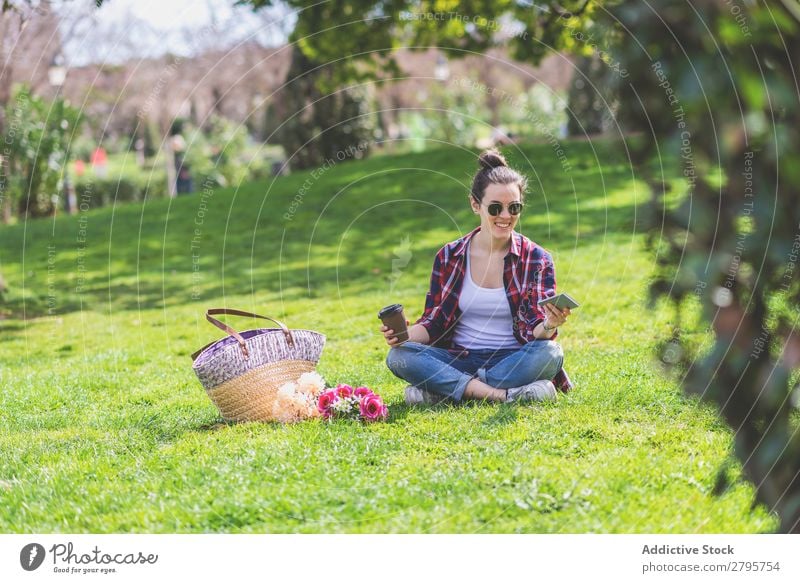 Vorderansicht einer jungen Hipsterfrau, die auf Gras in einem Park sitzt. Junge Frau Schickimicki trendy sitzen benutzend Boden Handy Lächeln Erholung