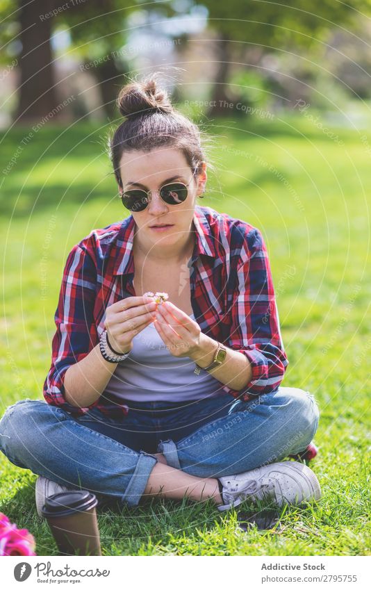 Vorderansicht einer jungen Hipsterfrau, die auf Gras in einem Park sitzt und an einem sonnigen Tag eine Blume hält. Junge Frau Schickimicki trendy sitzen Halt