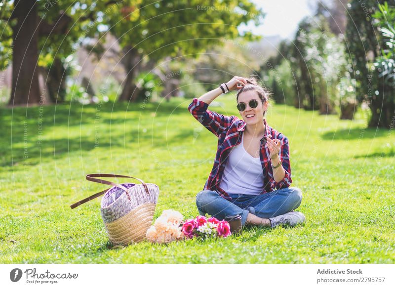 Vorderansicht einer jungen Hipsterfrau, die auf Gras in einem Park sitzt, während sie eine Blume hält und an einem sonnigen Tag lächelt. Frau Junge Frau