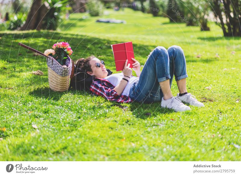 Seitenansicht einer glücklichen Frau, die am sonnigen Tag im Park auf Gras liegt, während sie ein rotes Buch liest. Profil Junge Frau Schickimicki trendy lügen