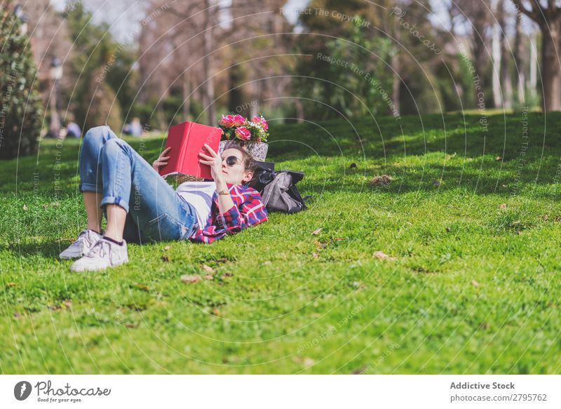 Seitenansicht einer glücklichen Frau, die am sonnigen Tag im Park auf Gras liegt, während sie ein rotes Buch liest. Profil Junge Frau Schickimicki trendy lügen